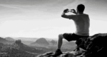 black and white photo of a man on a mountain top taking a photo with his Smart phone