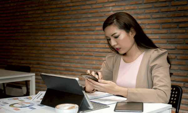 woman sitting at a table in front of a tablet and keyboard, using a cell phone